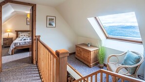 Ein gemütliches Schlafzimmer mit schrägen Wänden, einem hölzernen Bett, einem Nachttisch, einer Kommode, einem Korbstuhl und einem Fenster mit Blick auf eine verschneite Landschaft.
