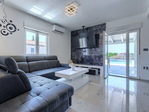 Ein modernes Wohnzimmer mit einem grauen Ecksofa, einem weißen Couchtisch, einem an einer Marmorwand montierten Fernseher und einer Glasschiebetür, die zu einem Pool führt.