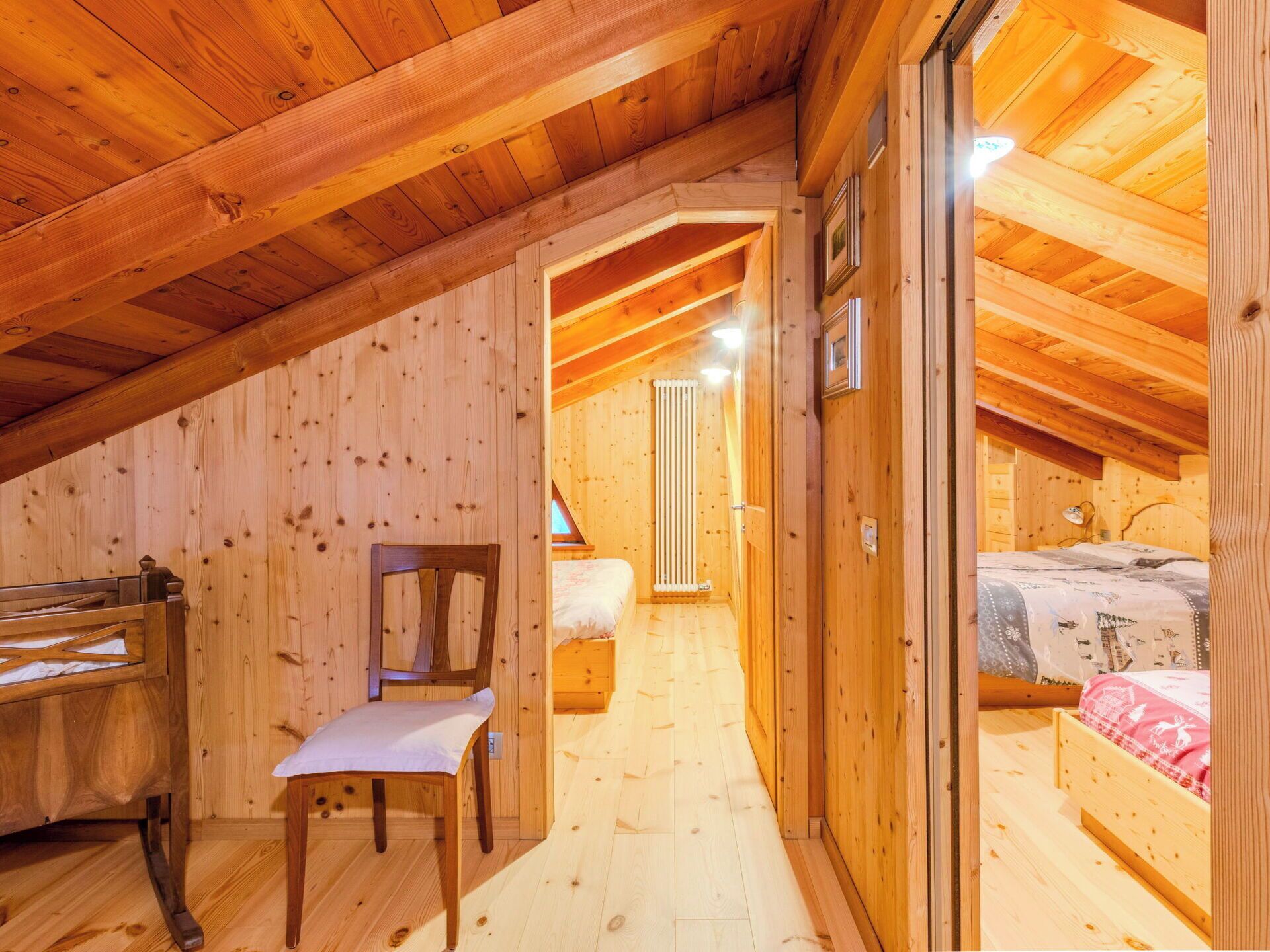 A wooden interior with a sloped ceiling, a wooden chair, a bed with a patterned bedspread, and a radiator.
