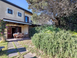 Ein zweistöckiges Haus mit einer Terrasse, die einen Tisch und Stühle bietet, umgeben von dichtem Buschwerk und einem großen Baum.