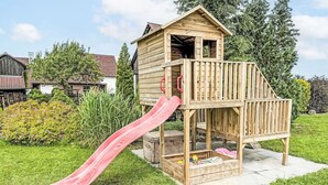 Een houten speelhuis met een glijbaan, omringd door een gazon en bomen.
