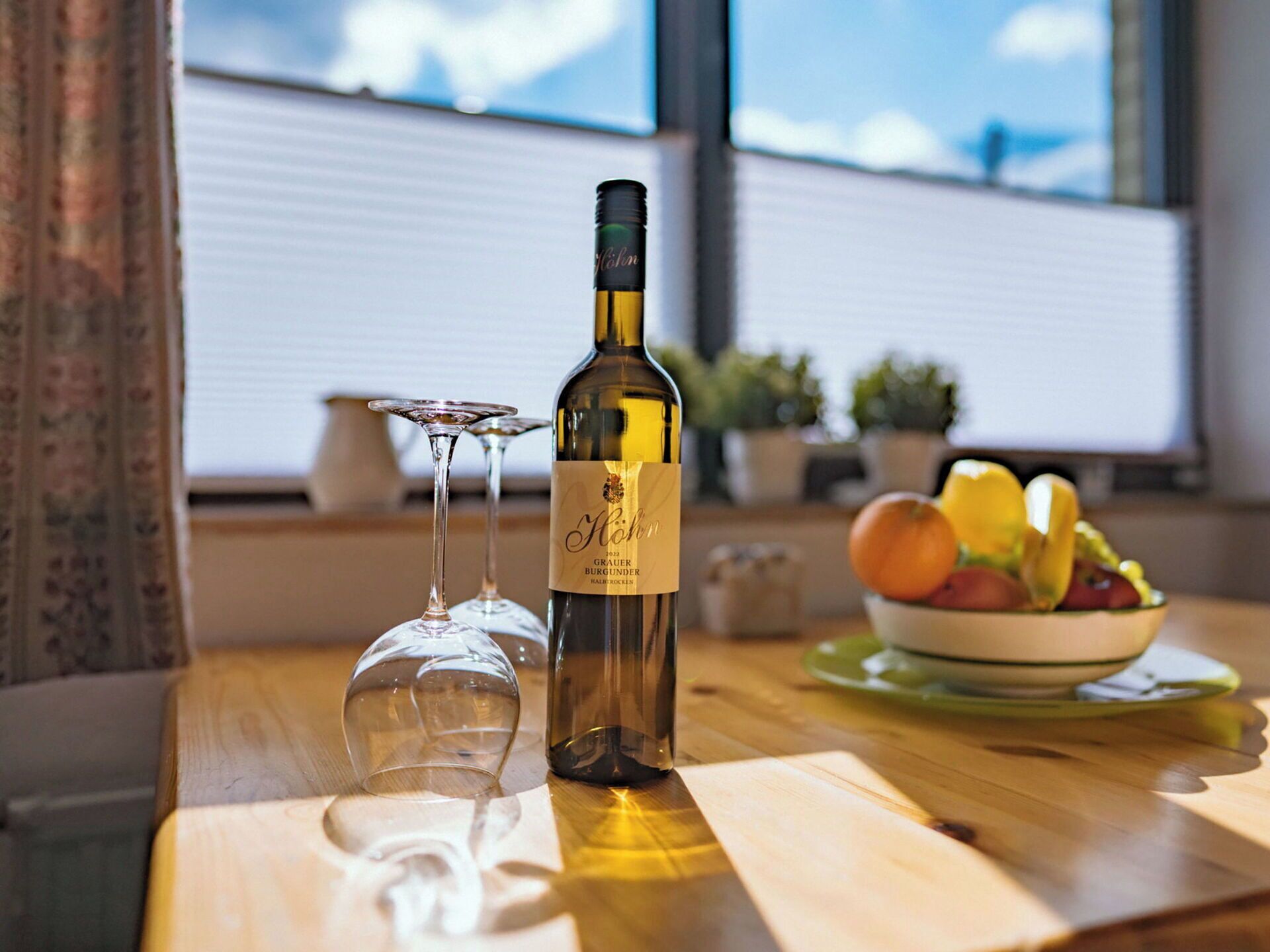 Une bouteille de vin et deux verres à vin sont posés sur une table en bois, avec un saladier rempli de fruits en arrière-plan.
