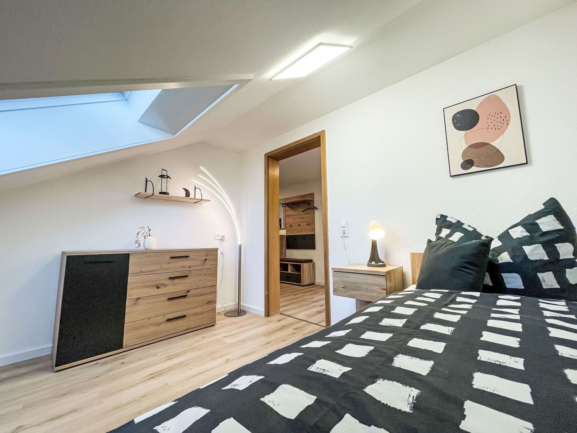 Een slaapkamer met dakraam, een bed met beddengoed met patroon, een nachtkastje met lamp en een aan de muur hangend kunstwerk.