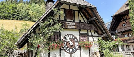 Un edificio tradicional de entramado de madera con un gran reloj, rodeado de vegetación y un cielo despejado.
