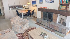 En stue med pejs, et spiseområde med stole, en sofa og et køkken i baggrunden.