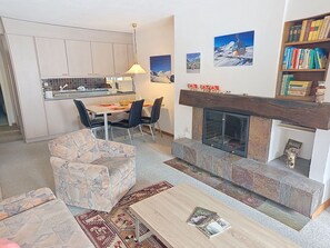 A living room with a fireplace, a dining area with chairs, a sofa, and a kitchen in the background. - Beautiful apartment for 4 guests with WIFI, pool, TV, balcony and panoramic view (Laax)