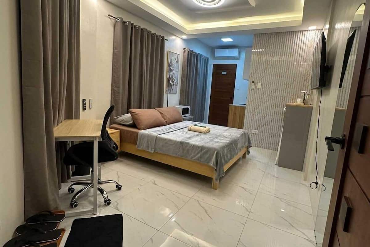 A modern bedroom with a bed, desk, and chair, a bathroom with a mirror and sink, and a TV mounted on the wall.