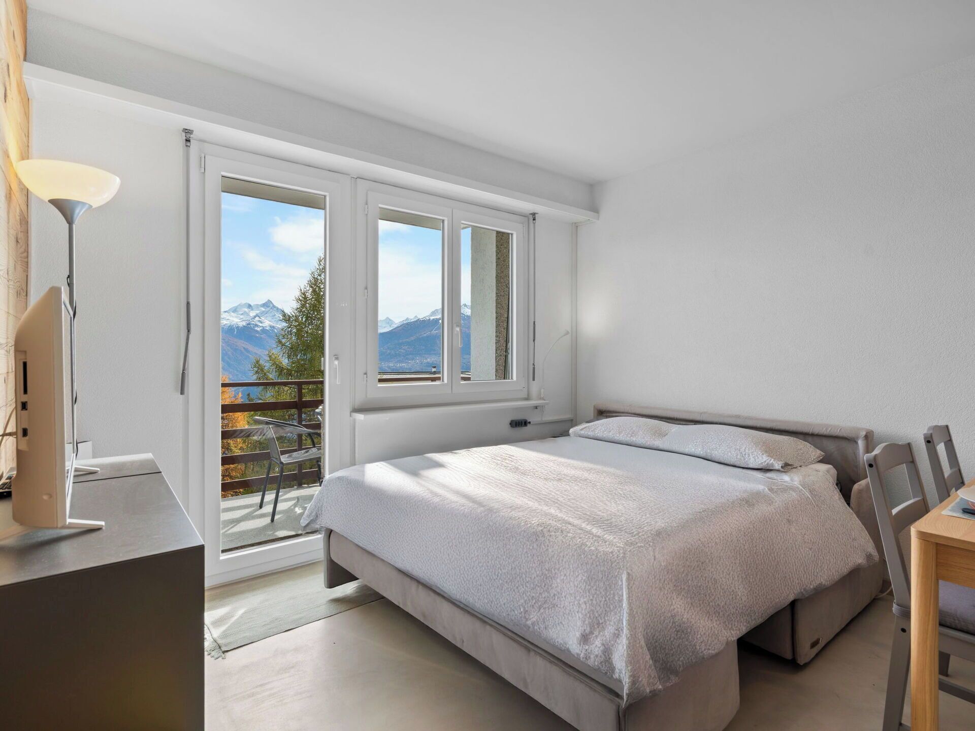 A bedroom with a bed, a desk, and a chair. There is a window with a view of mountains.