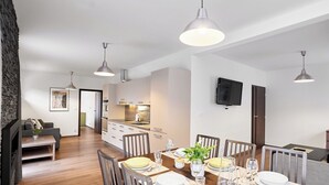 A modern dining area with a wooden table set for six, surrounded by chairs. A kitchen with white cabinets and a flat-screen TV is visible in the background.