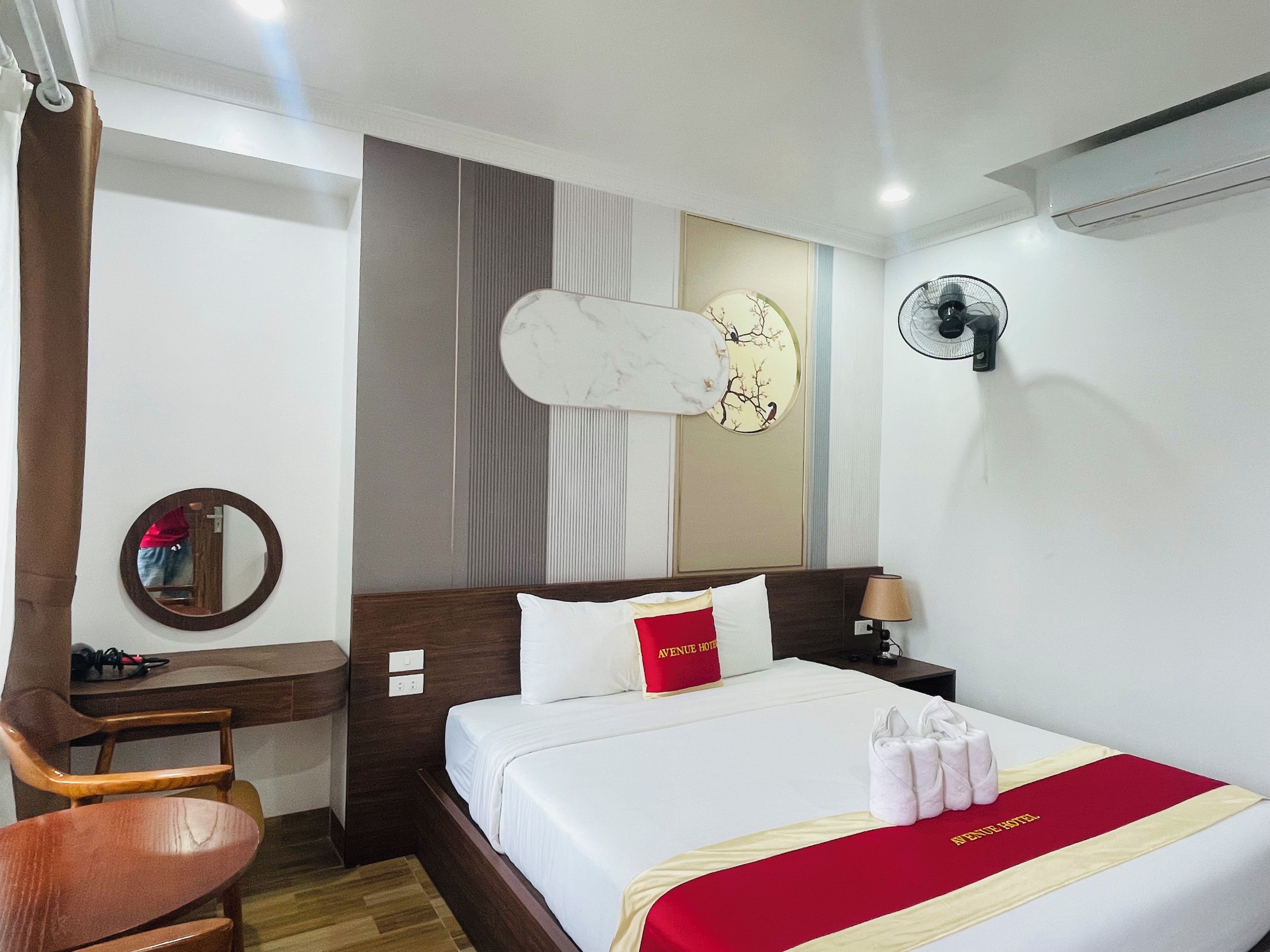 Une chambre d’hôtel comprenant un lit, un bureau, une chaise, un miroir, un ventilateur fixé au mur et un élément décoratif mural.
