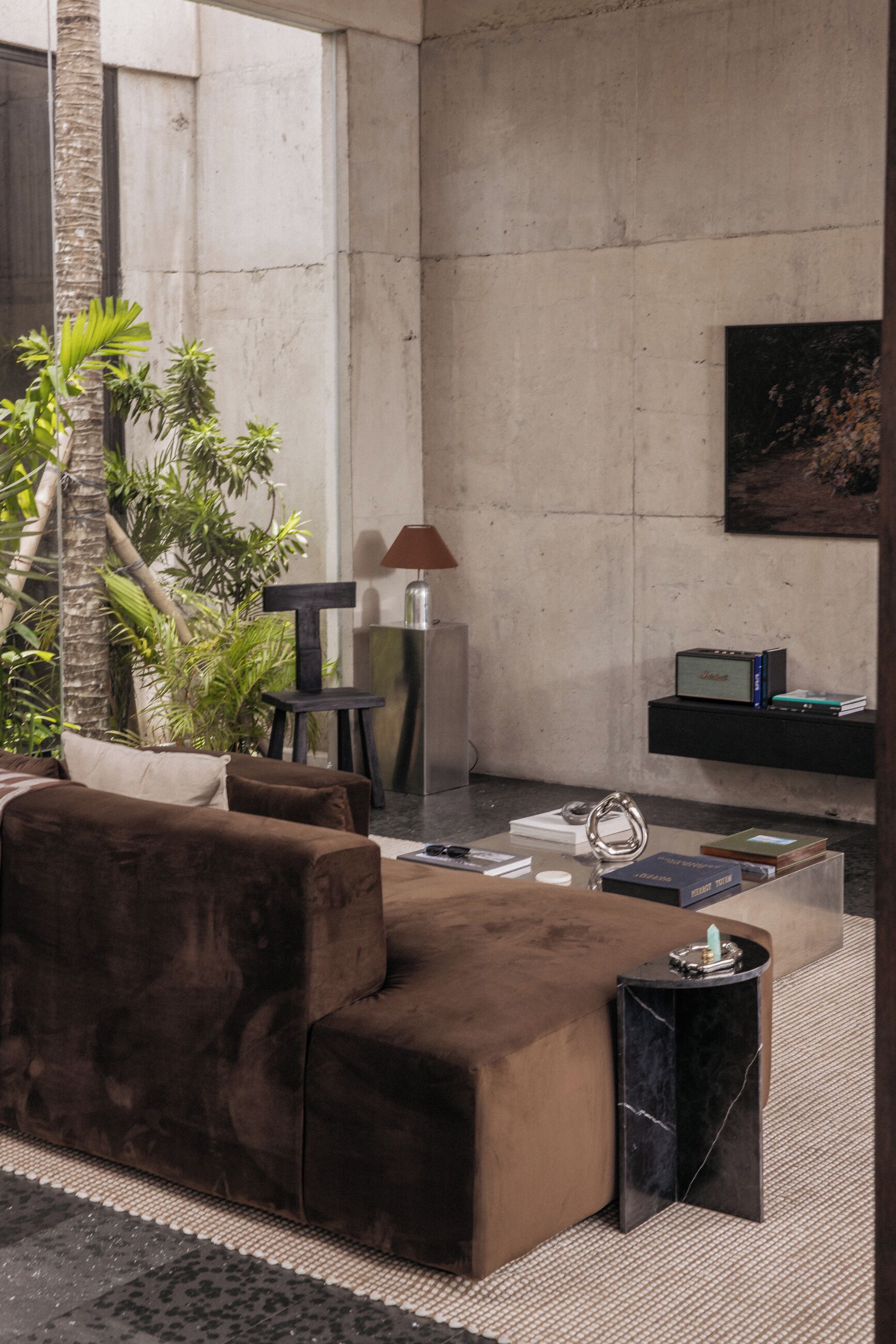 Un salon moderne avec un canapé marron, une petite table d’appoint, une lampe, une bibliothèque et une grande fenêtre donnant sur un paysage verdoyant.