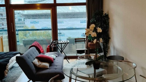 Een moderne woonkamer met een glazen eettafel, een bank en uitzicht op een bouwplaats door grote ramen.