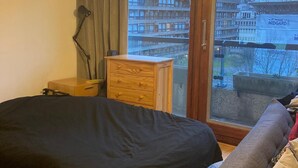 Een slaapkamer met een bed, nachtkastje, dressoir, lamp en uitzicht op een gebouw door een glazen deur.