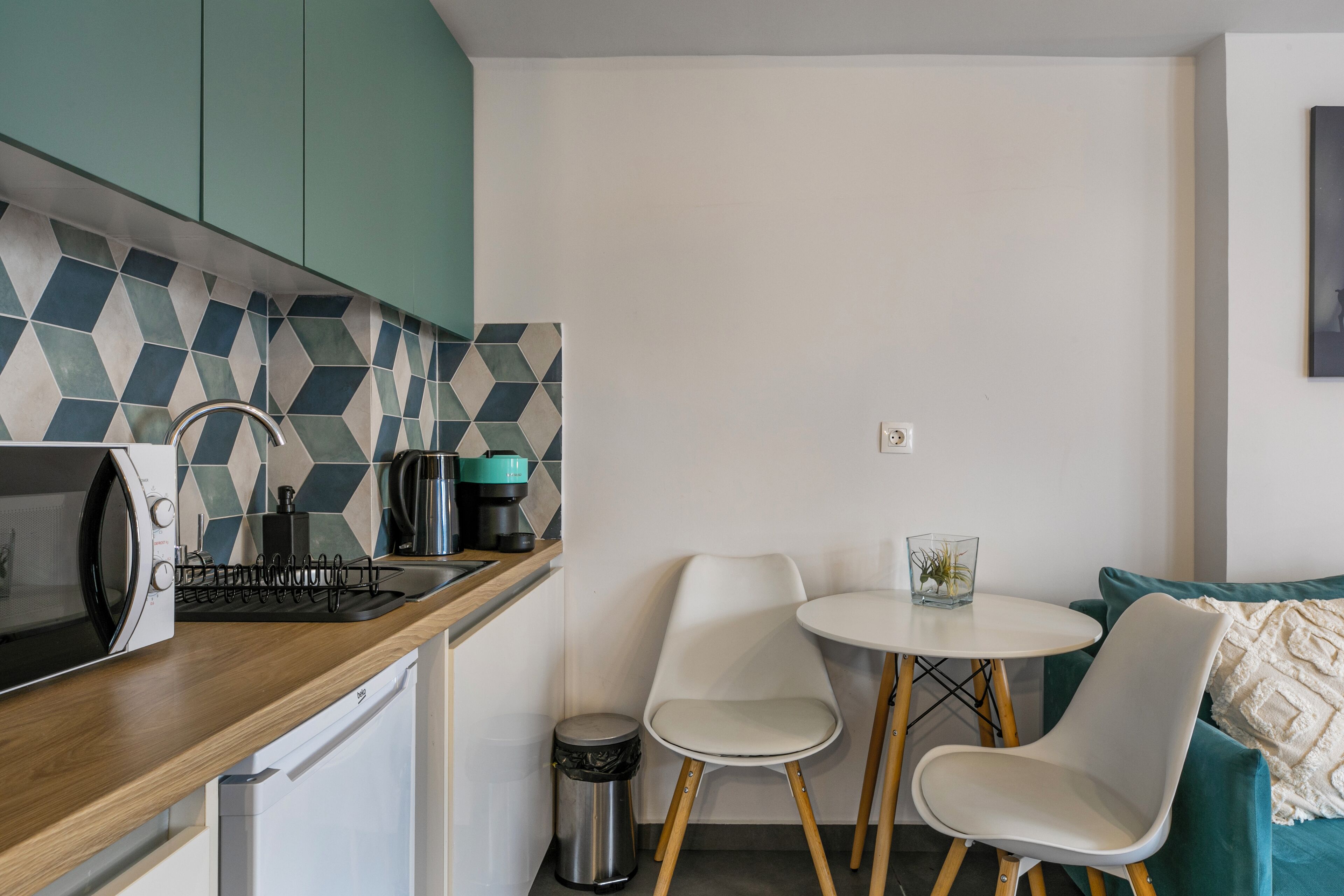 Ett kompakt kök med mikrovågsugn, kaffebryggare och diskho. Det finns två vita stolar och ett litet bord. Köket har turkosa skåp och ett kakel med geometriskt mönster bakom diskbänken.