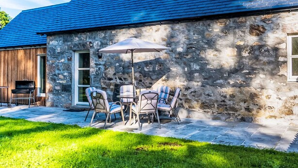 Un bâtiment en pierre avec un toit bleu, des sièges extérieurs avec des chaises et un parasol, et un barbecue sur la terrasse.