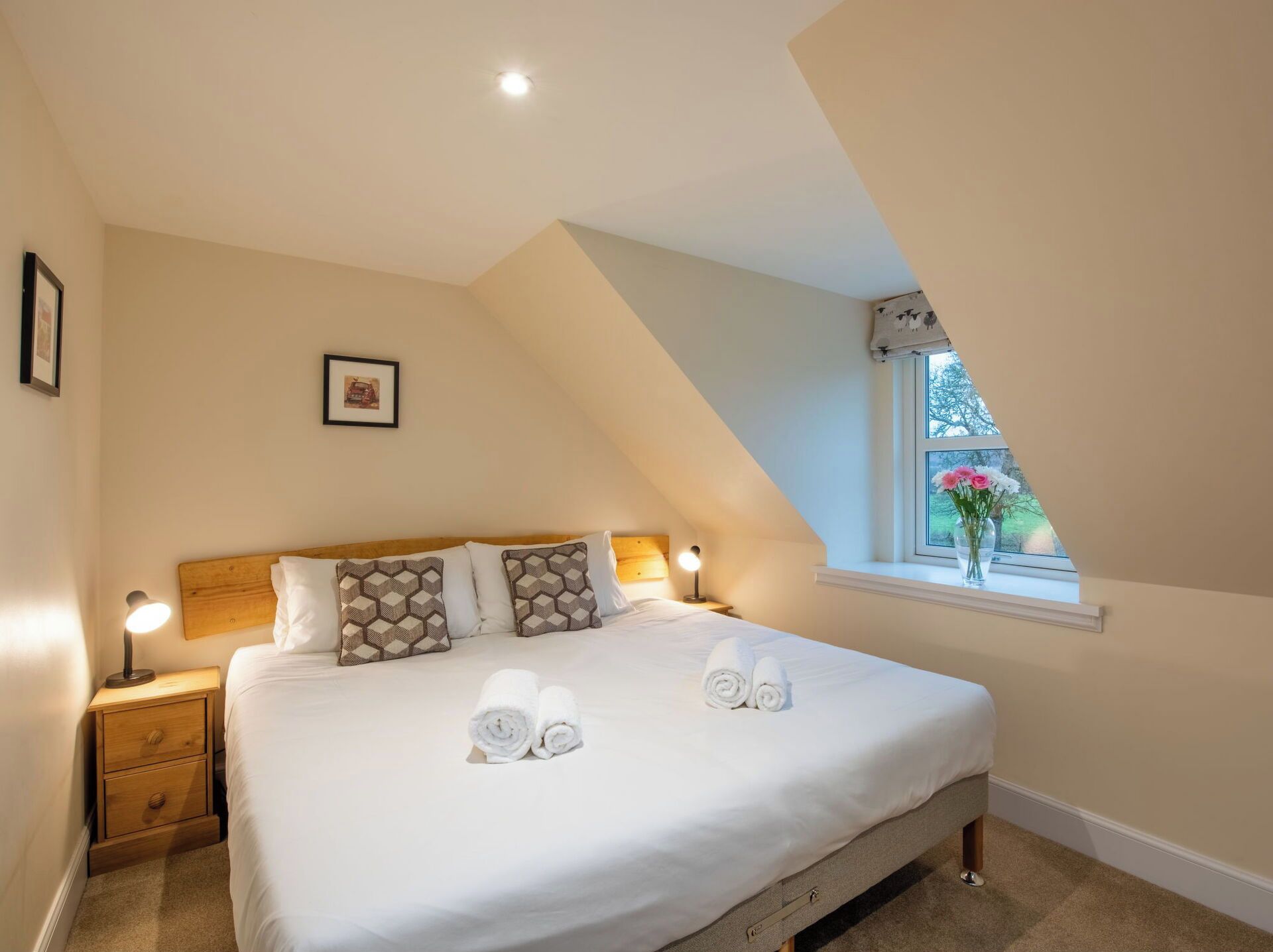 A bedroom with a wooden headboard, a bed with white linens and rolled towels, a nightstand with a lamp, and a window with a view of flowers.