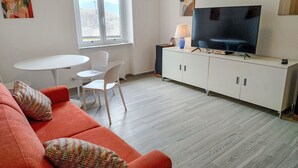 Una sala de estar moderna con un sofá rojo, sillas blancas, un televisor sobre un mueble blanco y un ventanal con vistas.