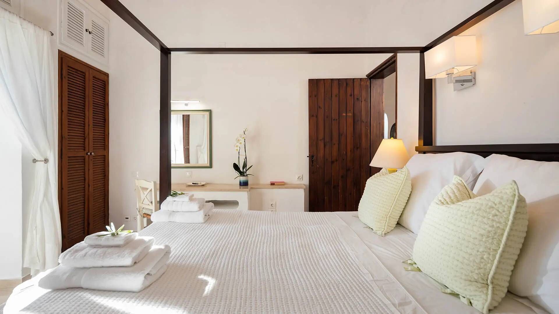 Una camera da letto con un letto grande, un comodino, uno specchio, un armadio in legno e un bagno visibile attraverso una porta aperta.