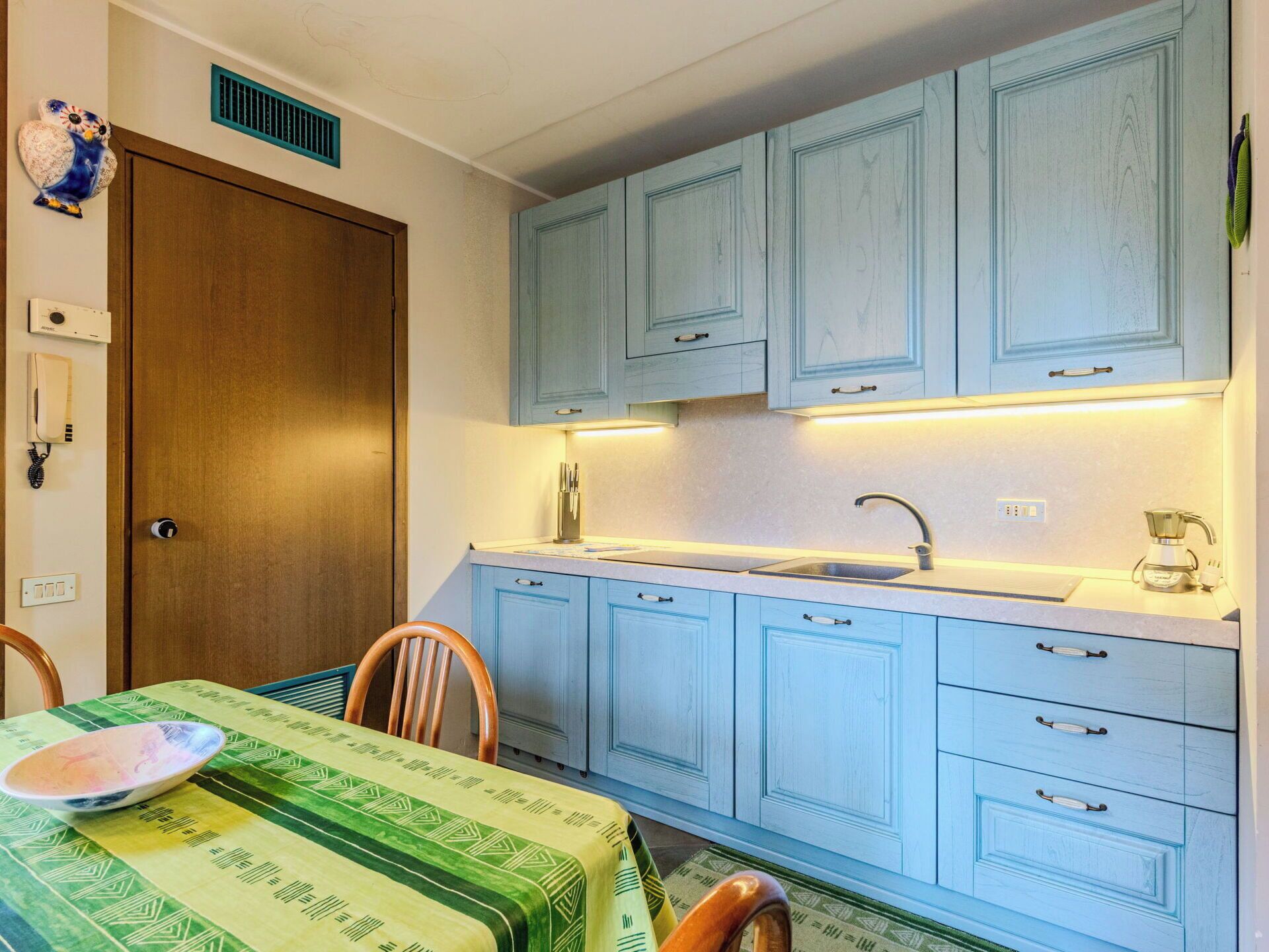 Une cuisine de taille réduite avec des meubles de couleur bleu clair, une porte en bois et une table recouverte d’une nappe à motifs verts.