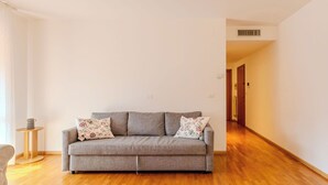 Un canapé de couleur neutre agrémenté de coussins à motifs floraux, dans une pièce dotée d’un plancher en bois et de murs blancs.