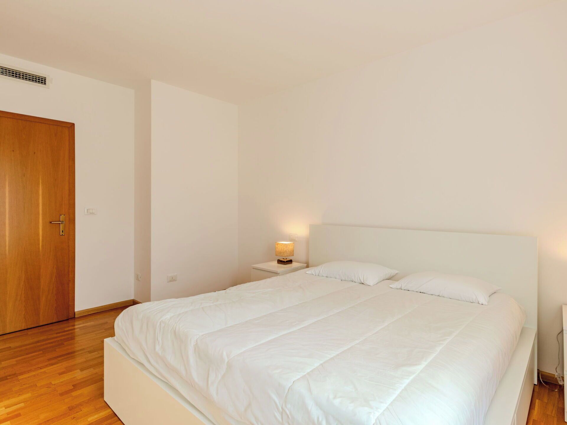Ein Schlafzimmer mit einem Bett, einem Nachttisch mit Lampe, einer Holztür und einer weißen Wand.