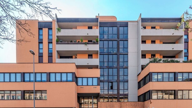 Un immeuble à plusieurs étages doté d’une architecture moderne, avec des balcons ornés de plantes, de grandes fenêtres et une entrée au niveau du sol, ainsi qu’un espace de stationnement pour vélos.