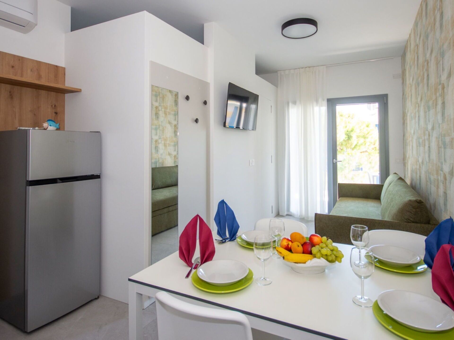 Une cuisine compacte équipée d’un réfrigérateur, d’une table à manger avec assiettes et verres, et d’un panier à fruits.