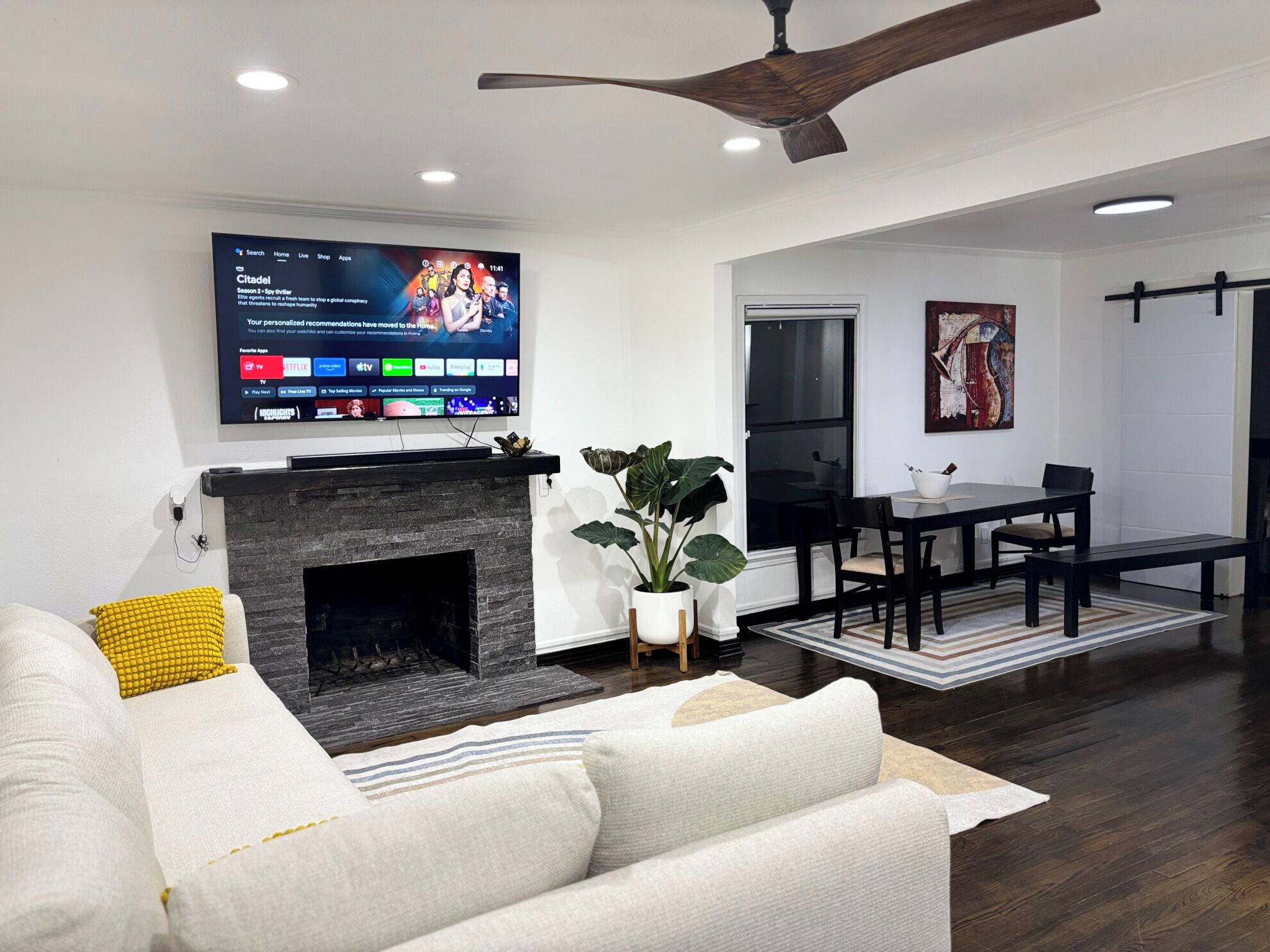 A modern living room with a flat-screen TV mounted above a fireplace, a dining area with a table and chairs, and a large potted plant.