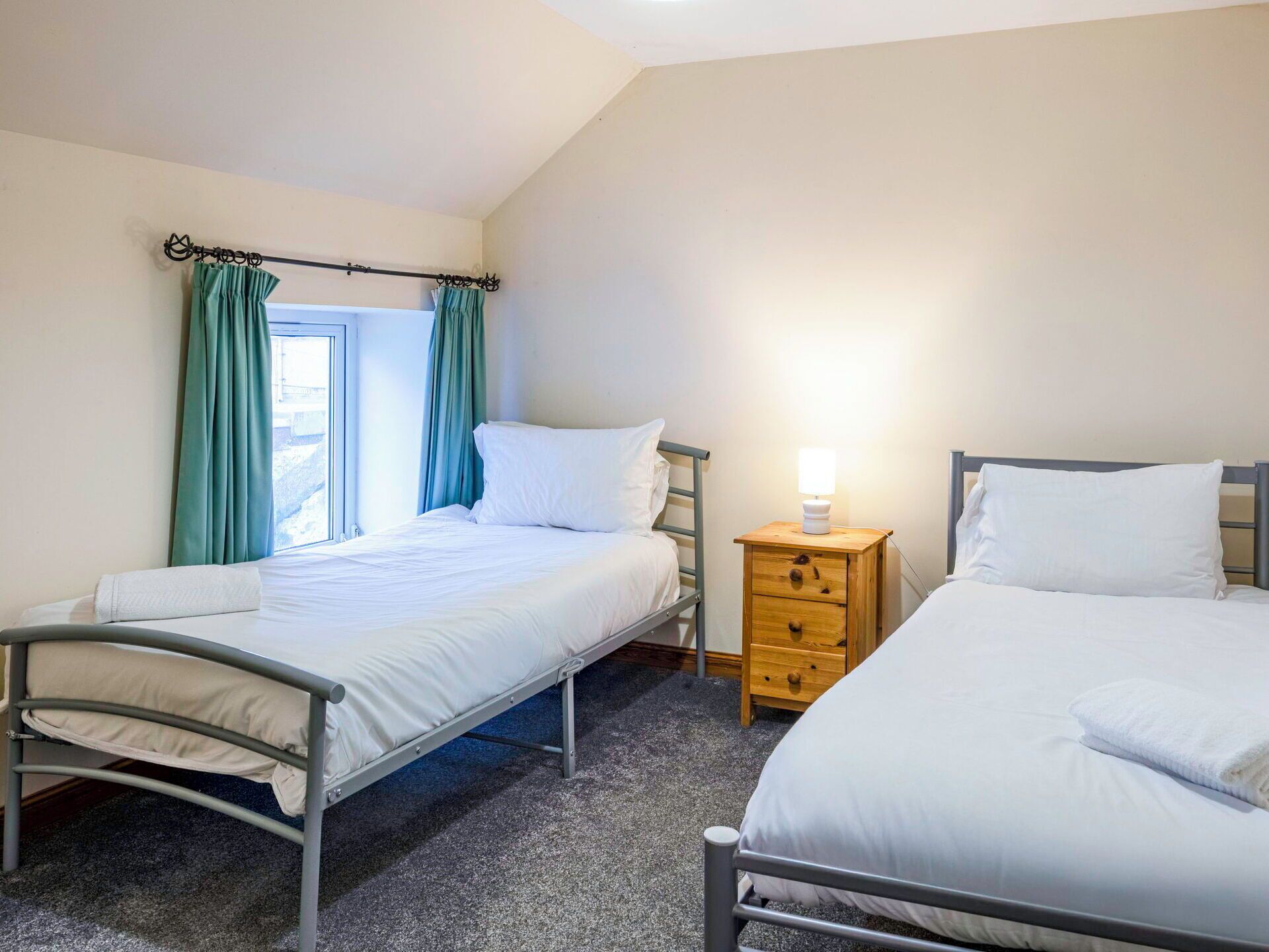 A bedroom with two beds, a window with curtains, a bedside table with a lamp, and a carpeted floor.