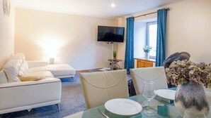Una habitación de hotel moderna con sofá, una mesa de comedor con platos y vasos, un televisor montado en la pared y una ventana con cortinas azules.