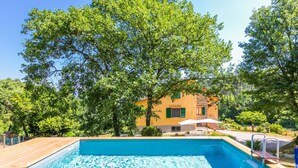 A swimming pool with a wooden deck, surrounded by lush greenery and a yellow building with a red roof. - Beautiful private villa for 10 people with A/C, WIFI, private pool and panoramic view (Vicchio)