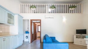 Un apartament modern amb cuina, zona d'estar amb un sofà blau, televisió i balcó amb plantes.