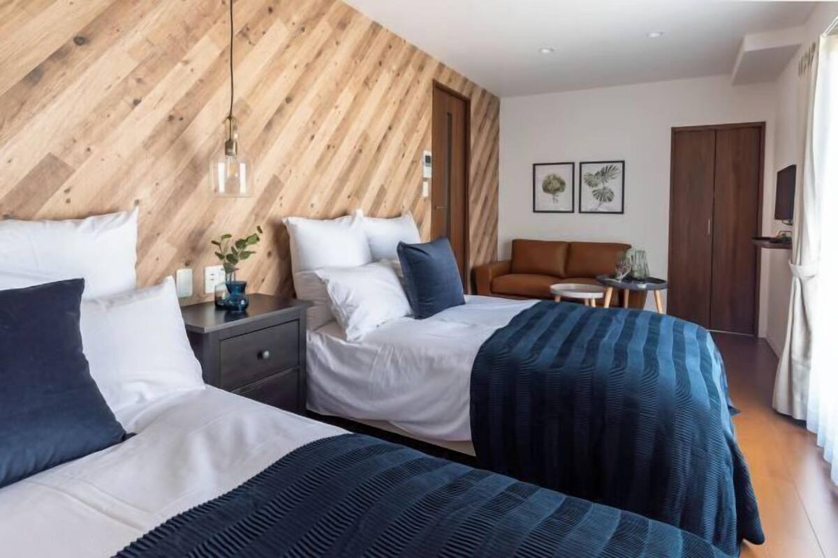 A hotel room with two beds, a wooden accent wall, a small table with a plant, and a TV on the wall.