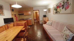 Una sala de estar acogedora con una mesa de comedor de madera, un sofá y un televisor. Se ve una cocina con armarios de madera y un lavamanos al fondo.