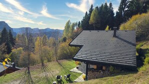 Ein Haus mit geneigtem Dach, einem Steinfundament und Blick auf Berge. Vor der Tür stehen zwei Stühle und ein kleiner Tisch.
