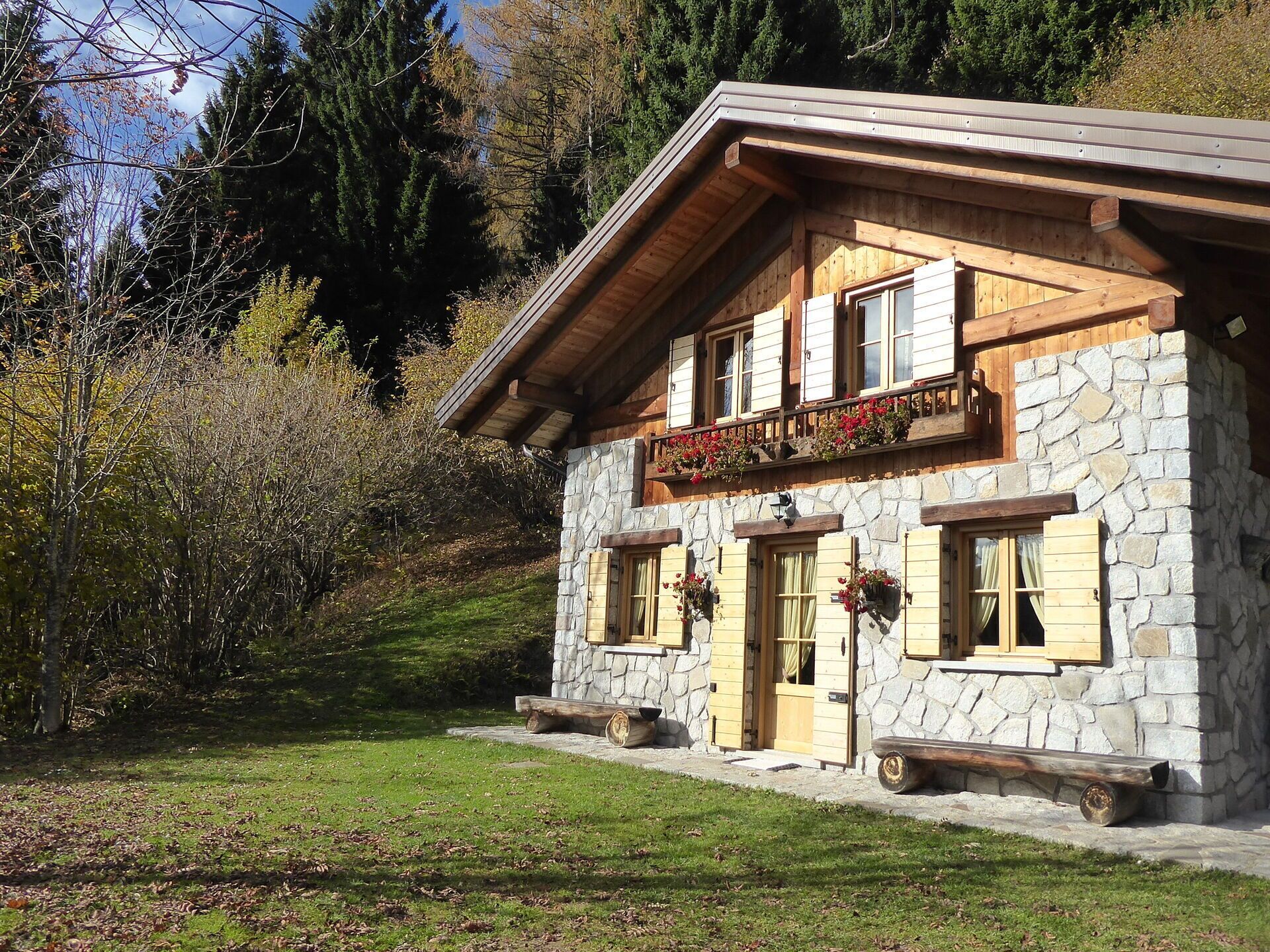 Un edificio de piedra con contraventanas de madera, un balcón lleno de flores y un banco rústico afuera.