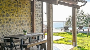 Een terras buiten met een houten tafel en bankjes, omgeven door stenen muren en uitzicht op zee.