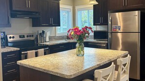 A modern kitchen with dark wood cabinets, a stainless steel refrigerator, a granite countertop island with white bar stools, pendant lighting, and a window with blinds. - The Nook at Latimer - 2 Bedroom Carriage House- Short Walk to Downtown and Beach (Penticton)