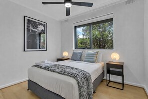 Un dormitorio con cama, dos mesitas de noche, ventilador de techo, ventana con vista a árboles y un cuadro en la pared.