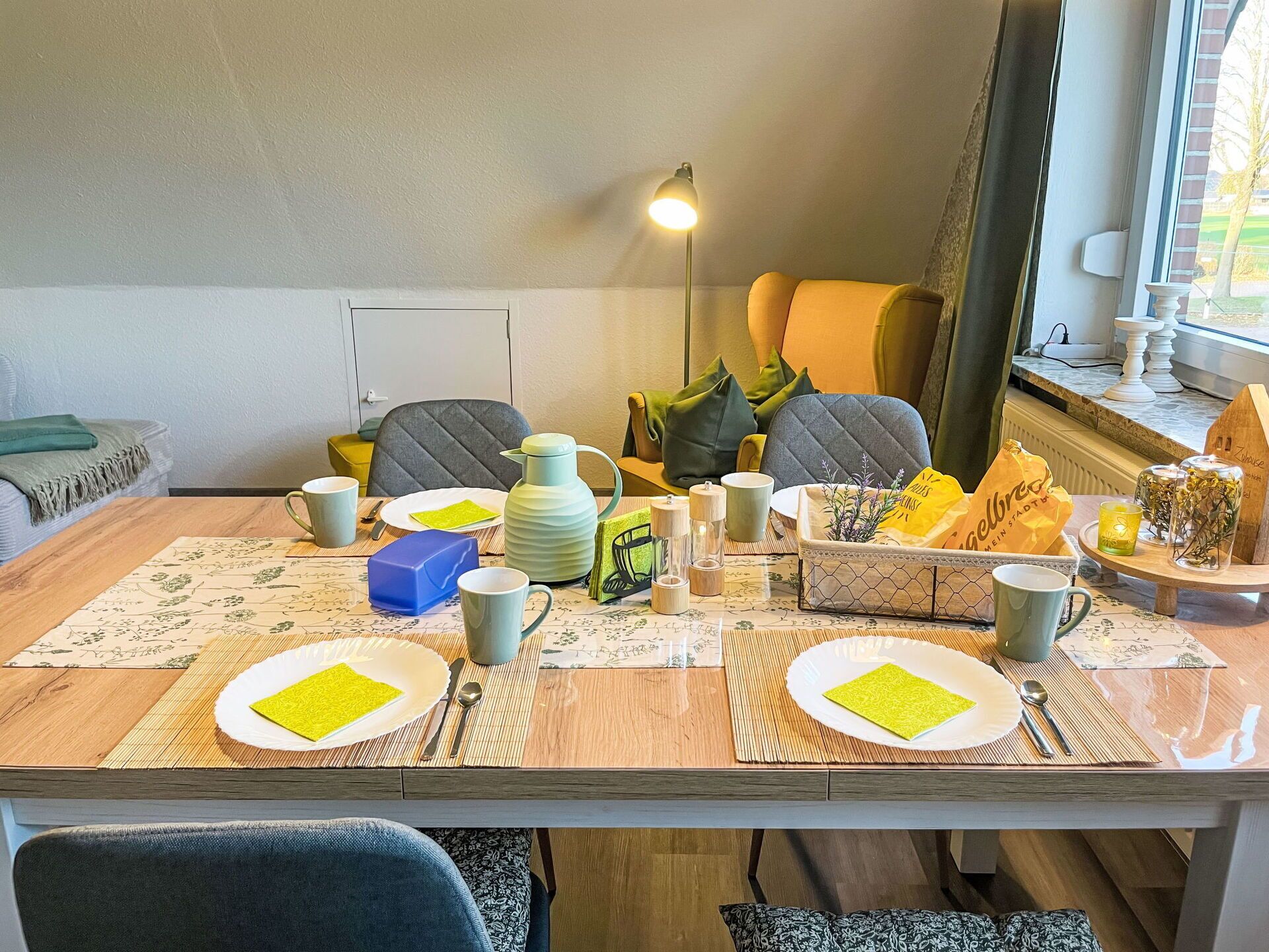 Un coin repas avec une table en bois dressée pour quatre personnes, comprenant des assiettes, des tasses et un pichet. Des chaises sont disposées autour de la table, et une lampe est visible à l’arrière-plan.