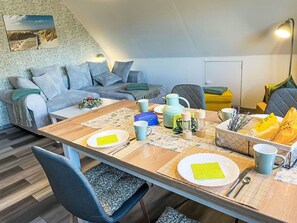 Un comedor con una mesa de madera dispuesta para cuatro personas, rodeada de sillas azules. Se observan un sofá azul y un sillón gris al fondo.