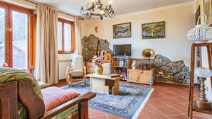 Een gezellige woonkamer met een houten bank, een salontafel, een televisie en een gitaar. Er is een groot raam met gordijnen, een kroonluchter en een vloerkleed.