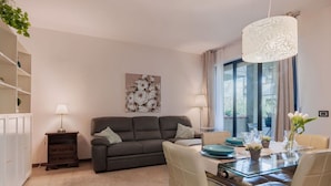 Una sala de estar moderna con un sofá, una mesa de comedor de cristal y sillas. Hay una lámpara, un cuadro en la pared y una ventana con cortinas.
