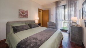 Un dormitorio con cama, una silla, una mesita de noche, una lámpara, un cuadro y una ventana con cortinas.