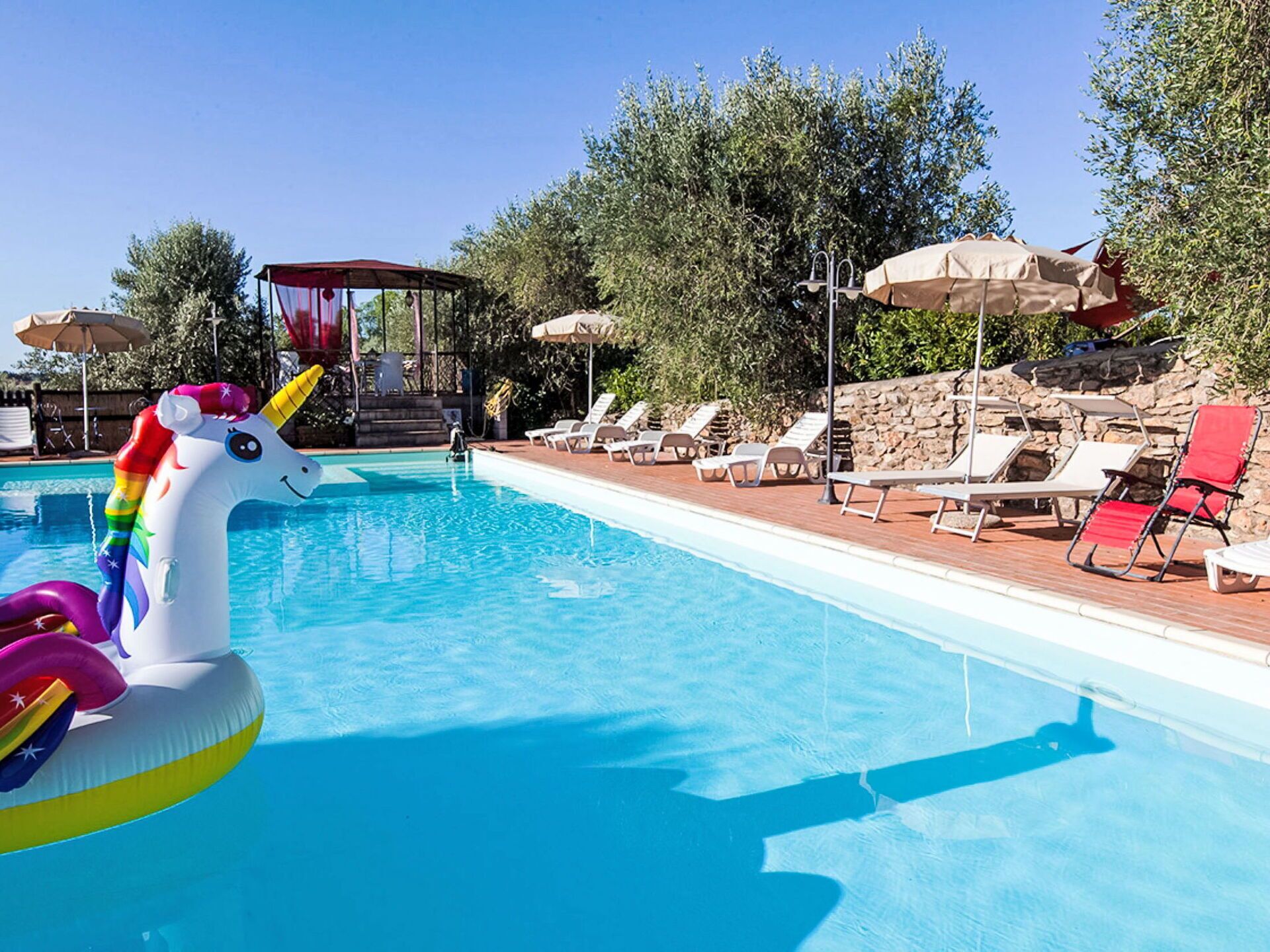 Una piscina con lettini e ombrelloni, circondata da alberi e un muro in pietra.