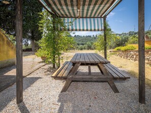 A wooden picnic table under a striped canopy, surrounded by gravel and greenery. - Nice apartment for 3 guests with A/C, WIFI, pool, terrace, pets allowed and panoramic view (Campiglia Marittima)