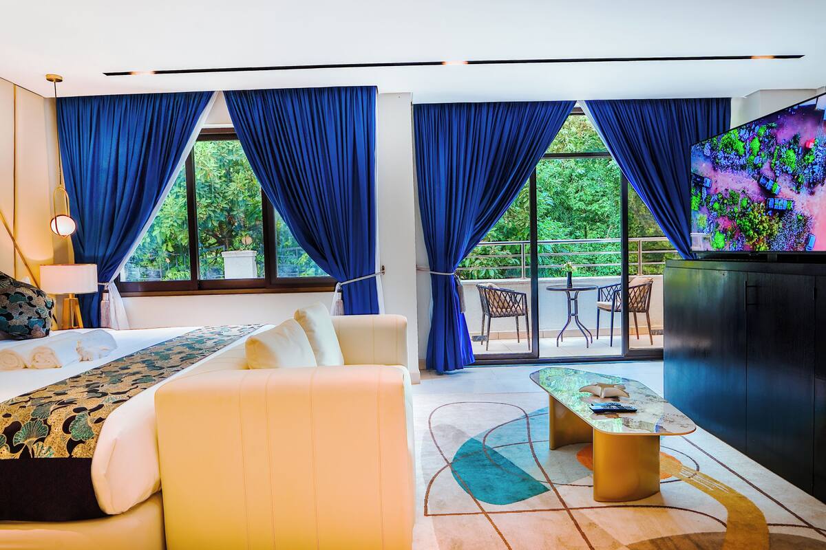 A modern hotel room with a sofa, a coffee table, a TV, and a balcony with blue curtains.