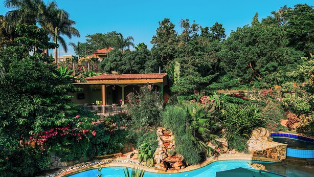 A pool surrounded by lush greenery and a building with a tiled roof. - Hotel Noesis  (Kampala)