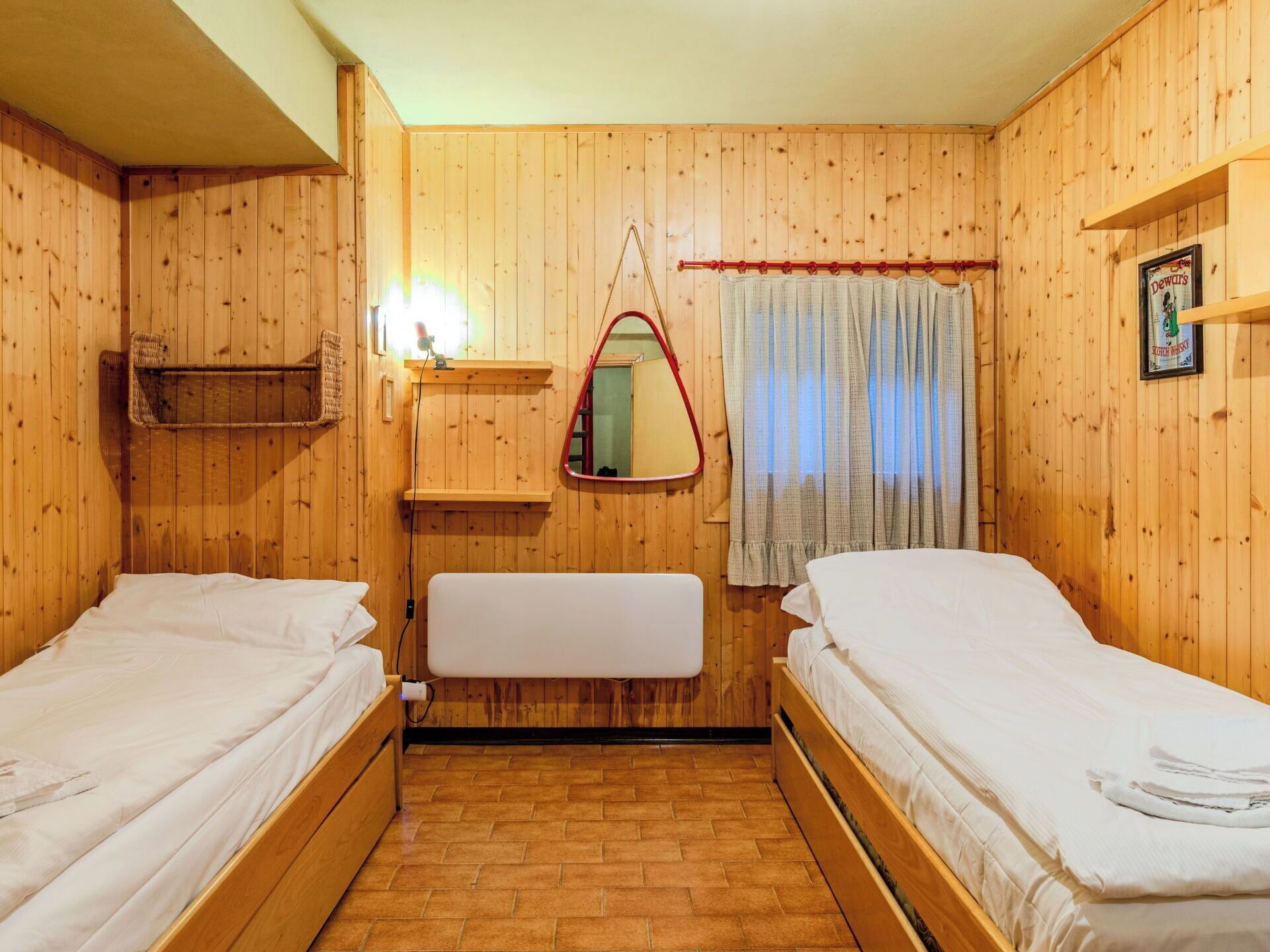 A wooden-paneled room with two single beds, a small shelf, a mirror, and a window with curtains.