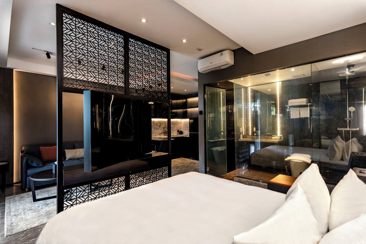Una habitación de hotel moderna con una cama grande, una zona de estar con un sofá y un sillón, televisión y cabina de ducha con paredes de vidrio.
